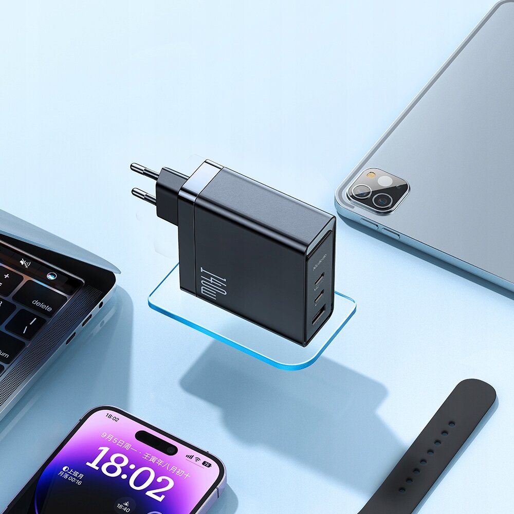 Na obrazie widoczna jest czarna ładowarka sieciowa z trzema portami USB, umieszczona na przezroczystej podstawce. W tle znajdują się laptop, smartfon z wyświetlaczem pokazującym godzinę 18:02 oraz tablet.