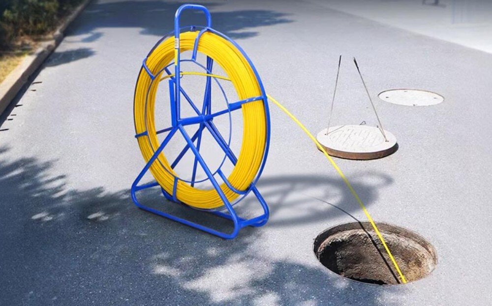 Pilot do przeciągania kabli EXTRALINK EX.31261 Niebieski stojak z żółtym przewodem ustawiony na asfalcie obok otwartego włazu kanalizacyjnego, do którego wprowadzony jest przewód