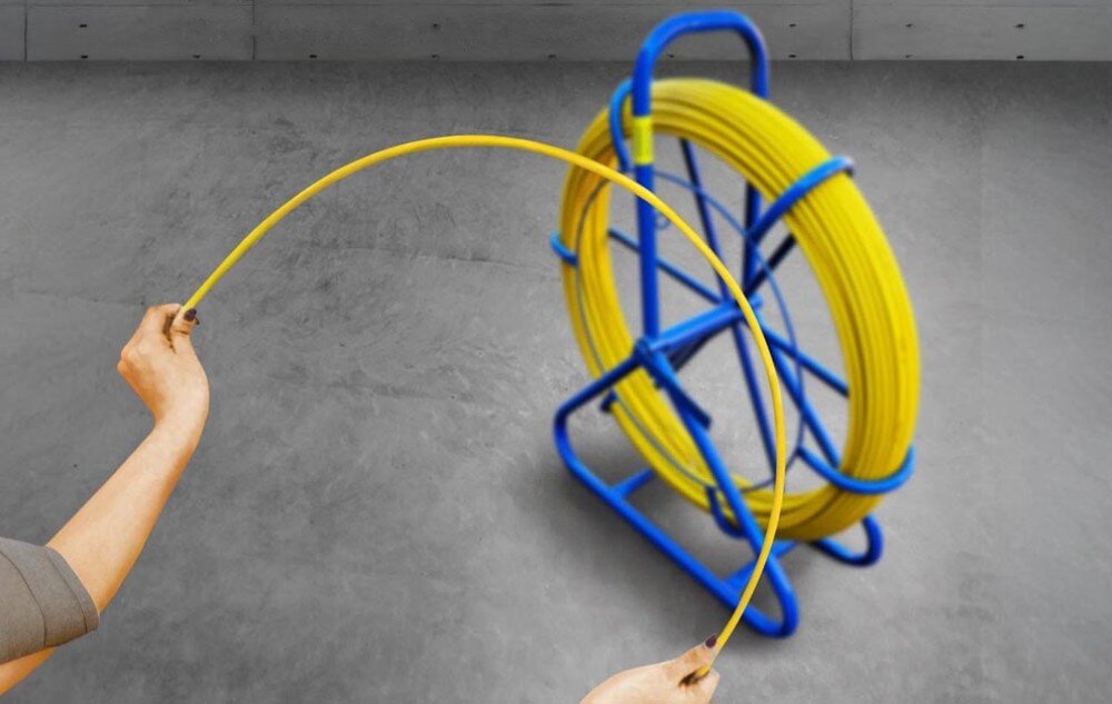 Pilot do przeciągania kabli EXTRALINK EX.31278 Dwie ręce zgięte w łuk trzymają żółty przewód, a w tle widać niebieski stojak z nawiniętą resztą przewodu na szarym podłożu