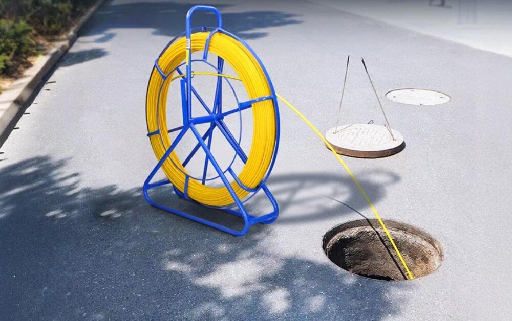 Pilot do przeciągania kabli EXTRALINK 30868 Niebieski stojak z żółtym przewodem ustawiony na asfalcie obok otwartego włazu kanalizacyjnego, do którego wprowadzony jest przewód