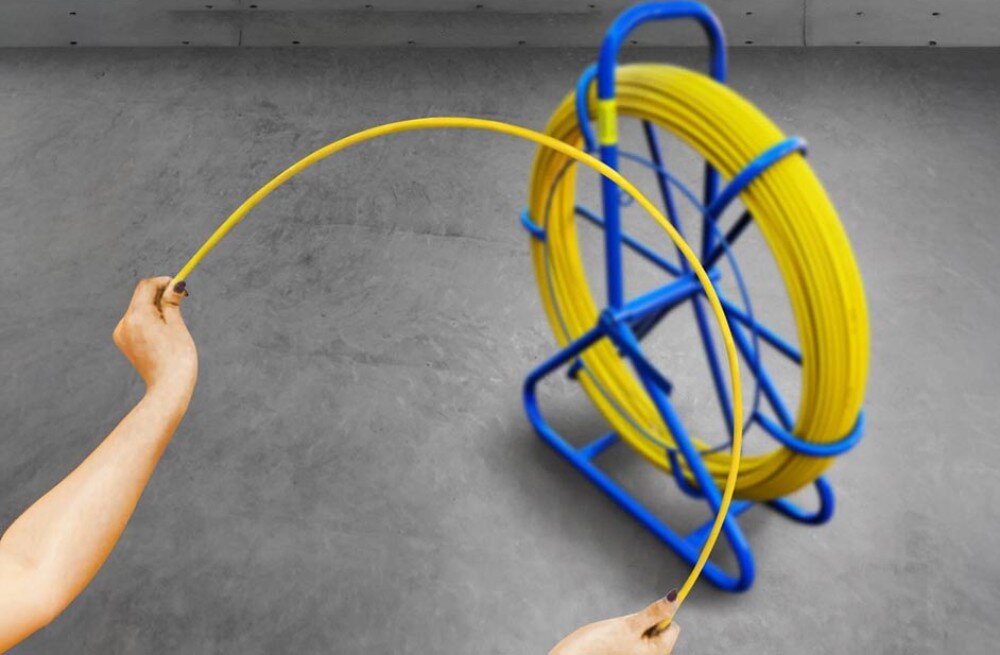 Pilot do przeciągania kabli EXTRALINK 30868 Dwie ręce zgięte w łuk trzymają żółty przewód, a w tle widać niebieski stojak z nawiniętą resztą przewodu na szarym podłożu