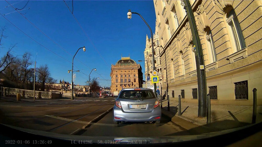 Zdjęcie przedstawia ulicę z zaparkowanym samochodem na poboczu, w tle widoczny jest zabytkowy budynek. Na dole obrazu widnieje napis: \'2022-01-06 13:26:49 14.41128E 50.0791N 35KM/H\'.