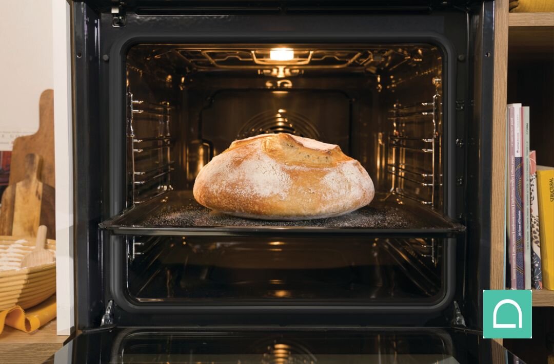 PIEKARNIK GORENJE BOS67371CLB Bochenek chleba piecze się na środkowym poziomie w otwartym piekarniku, oświetlonym ciepłym światłem od góry. Ujęcie eksponuje złocistą, popękaną skórkę oraz przestronne wnętrze komory