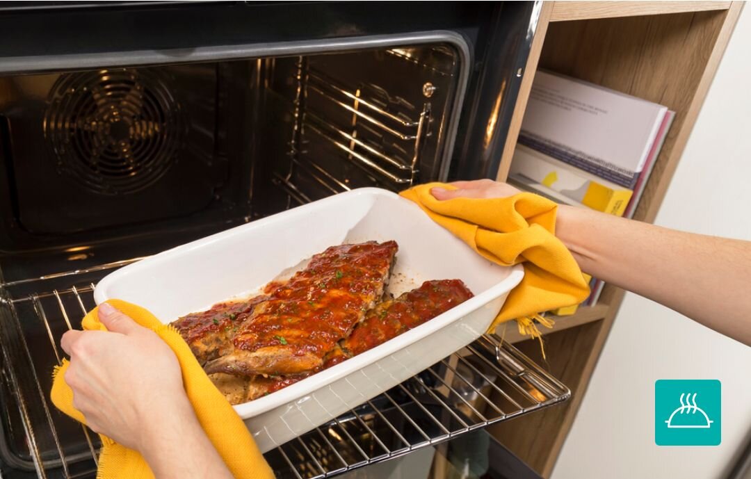 PIEKARNIK GORENJE BOS67371CLB Wyjmowanie gorącej potrawy w naczyniu żaroodpornym z piekarnika przy użyciu kuchennych rękawic. Zdjęcie akcentuje bezpieczeństwo i wygodę użytkowania