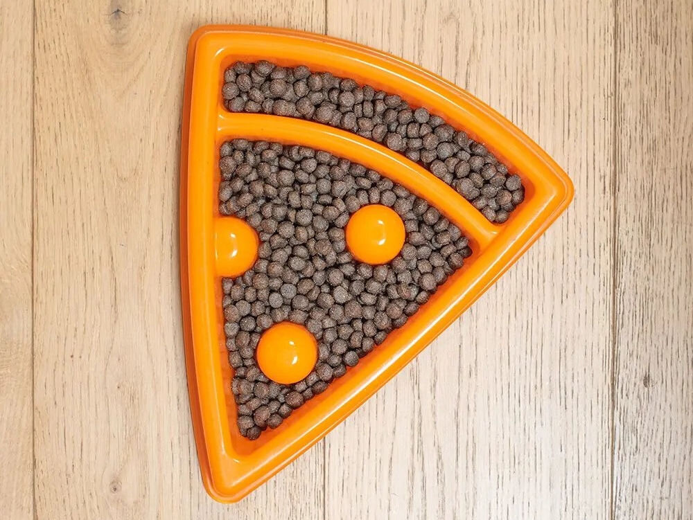 Miska spowalniająca dla psa ZIPPYPAWS Pizza ZP1415 Na zdjęciu znajduje się pomarańczowa miska w kształcie kawałka pizzy, wypełniona suchą karmą dla psa. Miska leży na drewnianej podłodze i ma trzy wypukłe elementy przypominające dodatki do pizzy