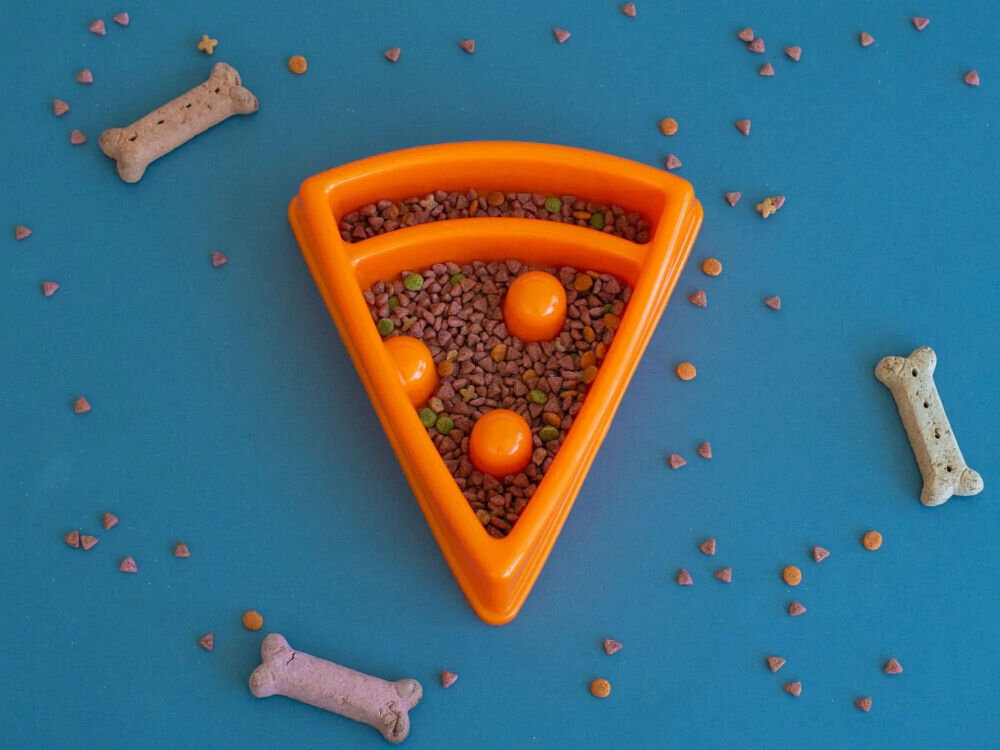 Miska spowalniająca dla psa ZIPPYPAWS Pizza ZP1415 Pomarańczowa miska w kształcie kawałka pizzy, wypełniona suchą karmą i trzema pomarańczowymi kulkami, leży na niebieskim tle, otoczona rozsypaną karmą i trzema psimi ciastkami w kształcie kości