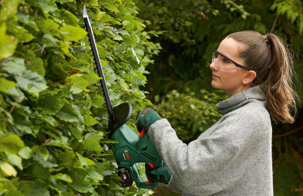 Nożyce do żywopłotu BOSCH Easy HedgeCut 18V-52-13 ogród osoba tnie żywopłoty trzymając nożyce w dłoniach w tle drzewa wskaźnik naładowania bieżąca kontrola energii do 170 m² żywopłotu na jednym cyklu ładowania czas ładowania około 124 minuty