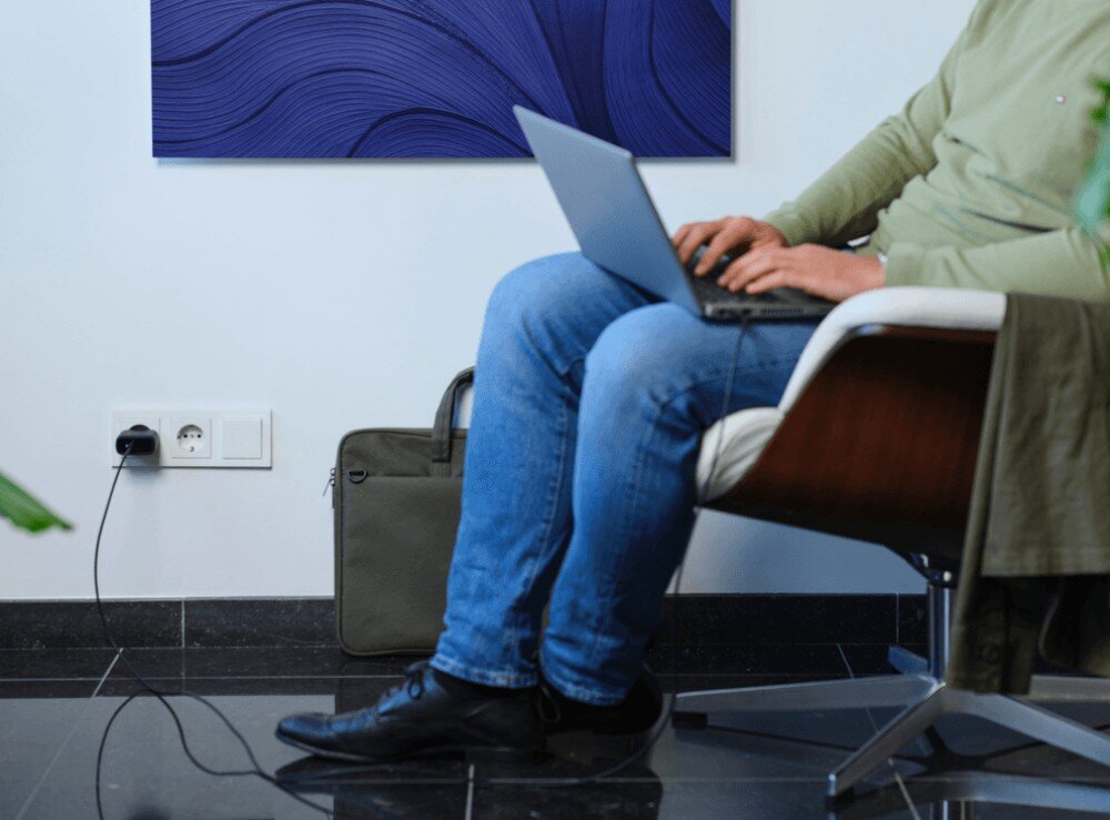 Zasilacz do laptopa TRUST Maxo 24817 65W Mężczyzna w jeansach siedzący na fotelu z laptopem na kolanach podłączonym długim kablem do ładowarki ściennej przy podłodze