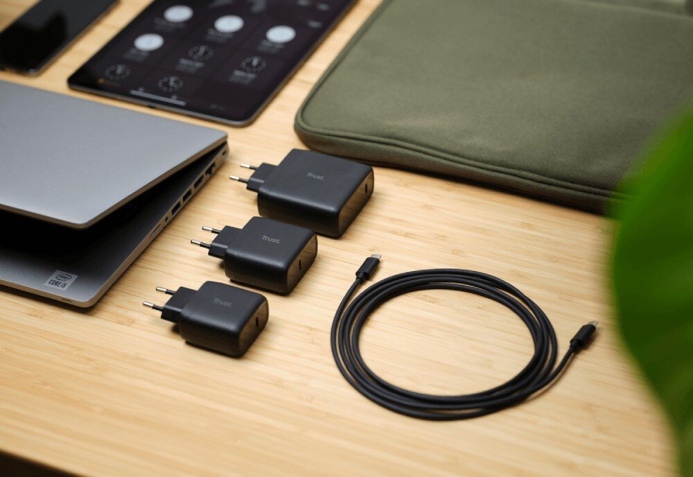 Zasilacz do laptopa TRUST Maxo 24817 65W Zestaw trzech czarnych ładowarek o różnej wielkości ułożonych obok siebie na drewnianym blacie wraz ze zwiniętym kablem USB-C i zamkniętym laptopem
