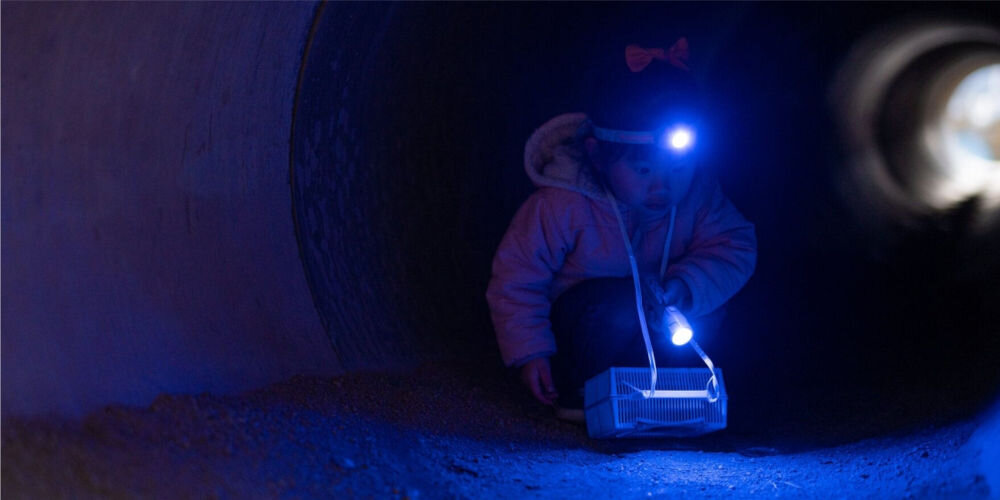 Latarka czołowa LEDLENSER KidLed4R Tęcza dziecko z latarką na czole siedzi w tunelu lampka świeci na niebiesko dwa tryby jasności Tryb niski 5 lumenów do 2 metrów czas pracy do 18 godzin Tryb standardowy 40 lumenów do 9 metrów do 2 godzin szybkie ładowanie około 60 minut praktyczne wygodne rozwiązanie