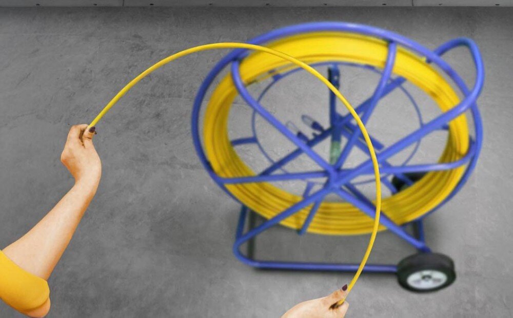 Pilot do przeciągania kabli EXTRALINK EX.15982 Dwie ręce zgięte w łuk trzymają żółty przewód, a w tle widać niebieski stojak z nawiniętą resztą przewodu na szarym podłożu