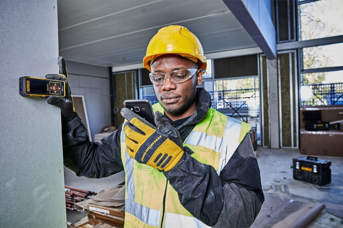 Dalmierz laserowy DEWALT DW03101-XJ 100m, pamięc, wewnętrzna, wartości, zapisywanie, mężczyzna spogląda na telefon trzymając dalmierz przy ścianie