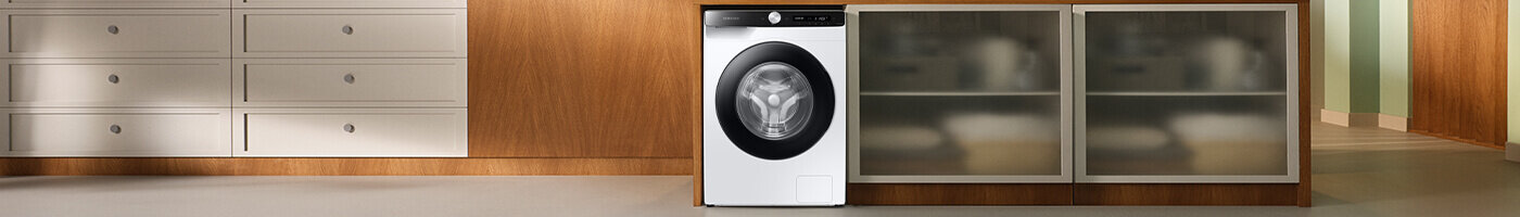 Na obrazie znajduje się pralka w zabudowie kuchennej, z białym frontem i czarnym panelem sterowania. Obok pralki widoczne są szafki z drewnianymi frontami i szufladami.