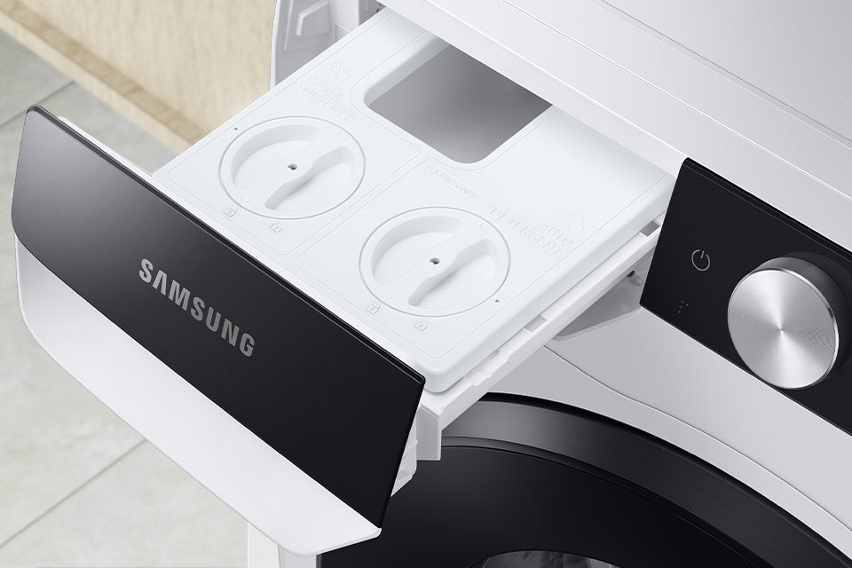 Zdjęcie przedstawia otwarty dozownik pralki z widocznymi dwoma okrągłymi pokrętłami. Na froncie dozownika znajduje się logo \'SAMSUNG\'.