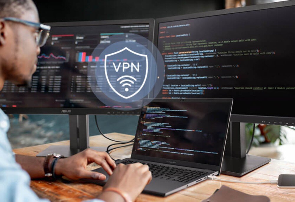 Router ASUS RT-AX88U Pro Mężczyzna siedzący przy biurku przed laptopem i dwoma monitorami z wyświetlonym kodem, na obrazie widoczna ikona z napisem VPN, połączenie VPN, szyfrowanie, protokoły, aplikacja