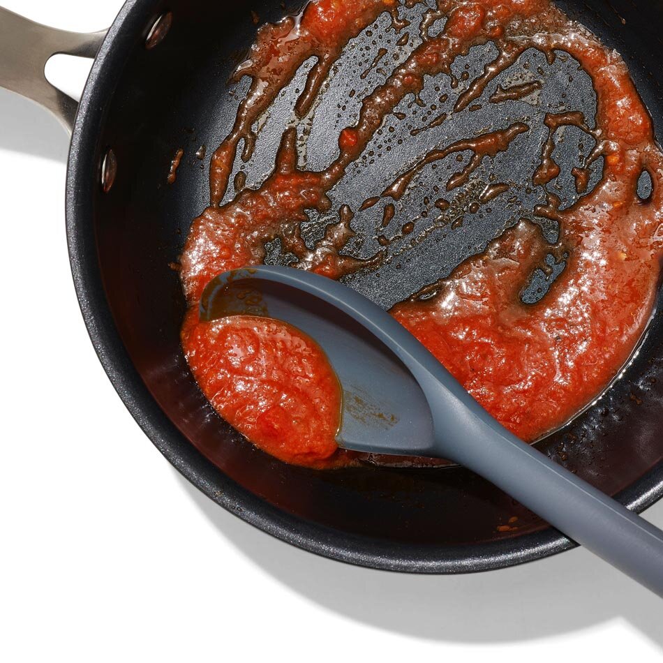Na obrazie widać czarną patelnię z resztkami czerwonego sosu oraz szarą silikonową łyżkę kuchenną. Łyżka znajduje się w patelni, nabierając część sosu.