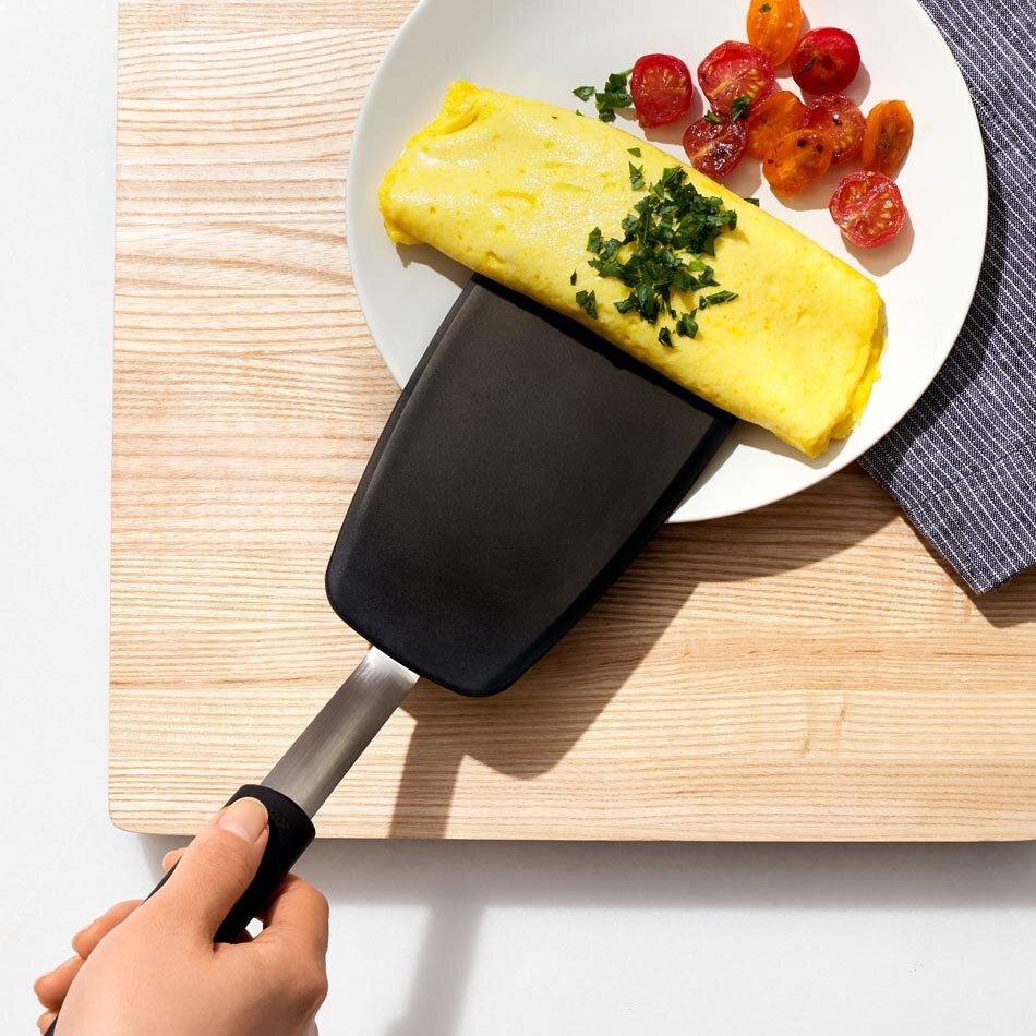 Na drewnianej desce znajduje się talerz z omletem i pokrojonymi pomidorami, a silikonowa łopatka podtrzymuje omlet. Dłoń trzyma uchwyt łopatki.