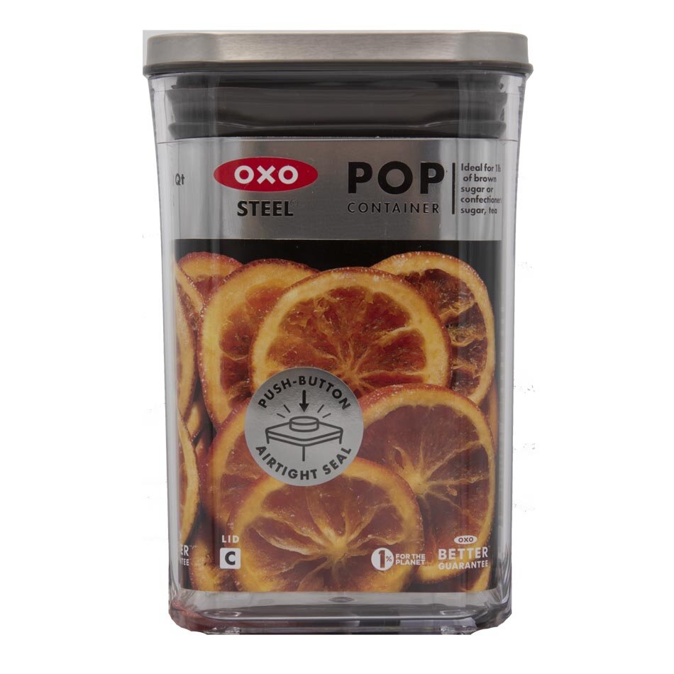 Kwadratowy pojemnik kuchenny z przezroczystego tworzywa z metalową pokrywką, ozdobiony grafiką suszonych pomarańczy. Na etykiecie widnieje napis: \'OXO STEEL POP CONTAINER\' oraz \'PUSH-BUTTON AIRTIGHT SEAL\'.