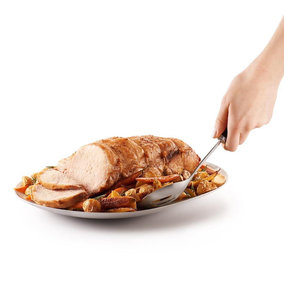 Na talerzu znajduje się pieczeń z mięsa otoczona pieczonymi warzywami. Ręka trzyma perforowaną łyżkę kuchenną, która dotyka jedzenia.
