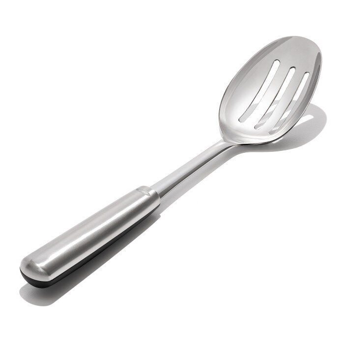 Na obrazie widoczna jest metalowa łyżka cedzakowa z trzema podłużnymi otworami na główce. Rączka jest gładka i błyszcząca, z widocznym napisem \'oxo\'.