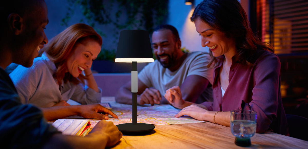 Lampa stołowa PHILIPS HUE 929003128401 Biały, szczelność, IP54, bateria, 48 godzin, 4 osoby siedzą przy stole z włączoną lampką wieczorową porą