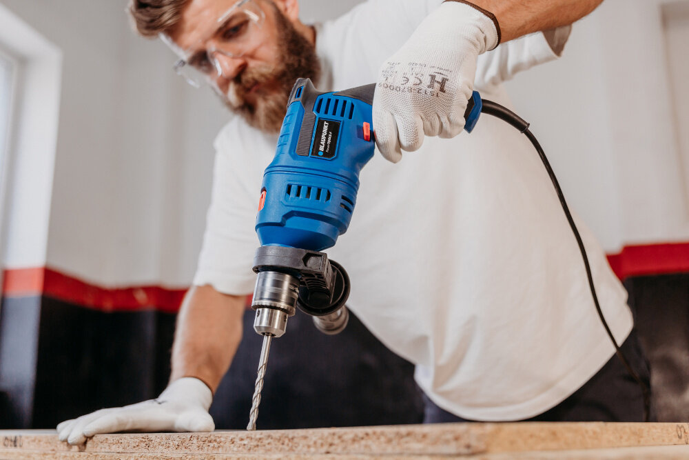 Wiertarka udarowa BLAUPUNKT ID8010 pomieszczenie drewniany blat osoba trzyma wiertarkę w dłoni wierci otwór w drewnianym elemencie w tle jasna ściana uniwersalne narzędzie do wiercenia w drewnie, betonie i stali moc 900 W moc znamionowa 710 W funkcja udaru regulacja prędkości 0–3000 obr./min