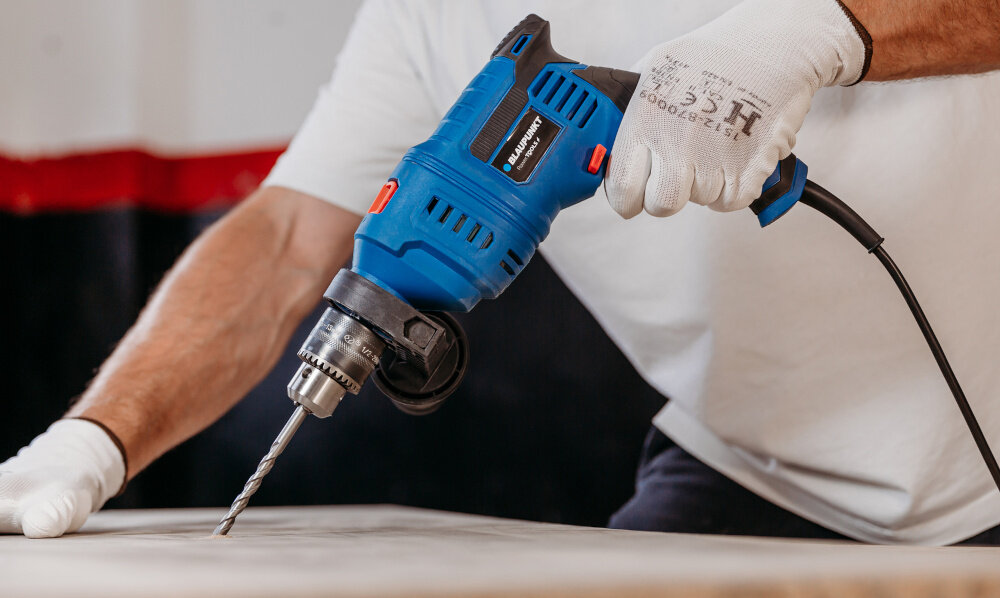 Wiertarka udarowa BLAUPUNKT ID8010 pomieszczenie drewniany blat osoba wierci otwór w blacie w tle jasna ściana funkcjonalność wygodne użytkowanie wiercenie otworów o średnicy do 25 mm w drewnie do 13 mm w betonie do 10 mm w stali wysoka precyzja działania mocny udar