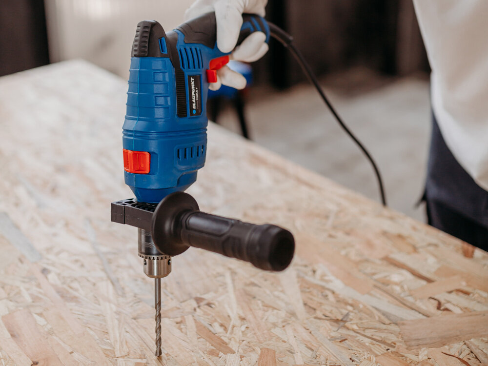 Wiertarka udarowa BLAUPUNKT ID8010 pomieszczenie drewniany blat osoba trzyma wiertarkę w dłoni wierci otwór w blacie uchwyt pomocniczy zwiększa stabilność prowadzenia poprawia bezpieczeństwo pracy główna rękojeść ma ergonomiczną konstrukcję ułatwia prowadzenie narzędzia ogranicza zmęczenie dłoni antypoślizgowa powłoka lepsza kontrola nad wiertarką
