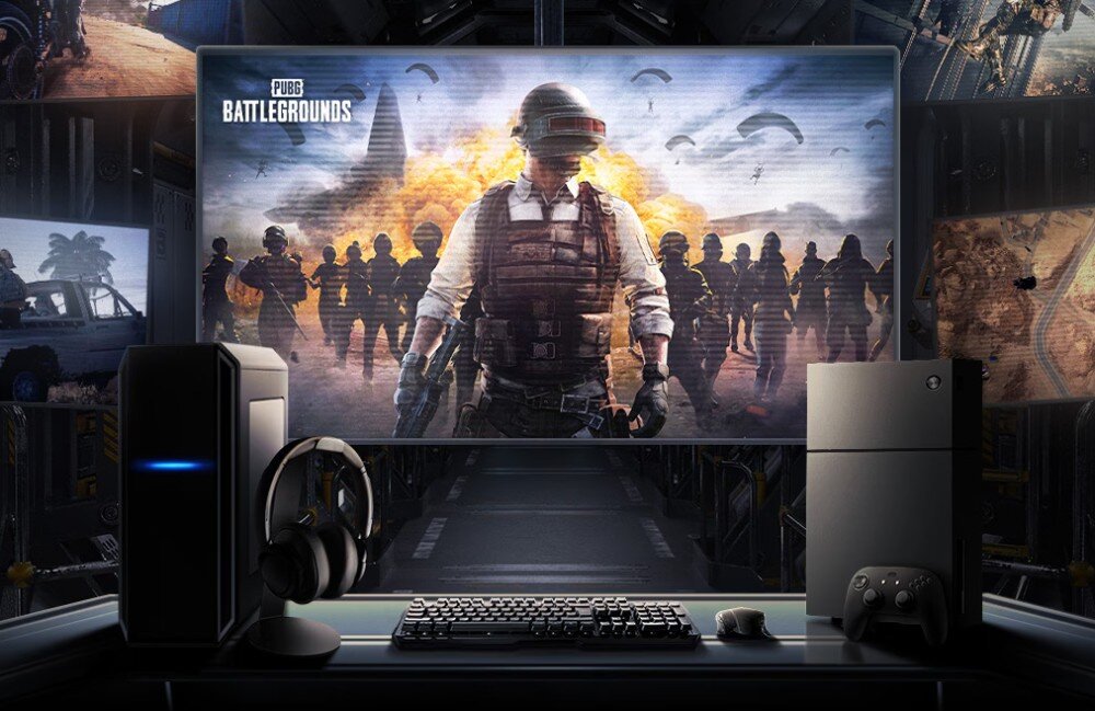 Na obrazie widoczny jest ekran z grą \'PUBG BATTLEGROUNDS\', przedstawiający postać w hełmie i grupę ludzi na tle eksplozji. Przed ekranem znajduje się zestaw komputerowy z klawiaturą, myszką, słuchawkami i konsolą do gier.