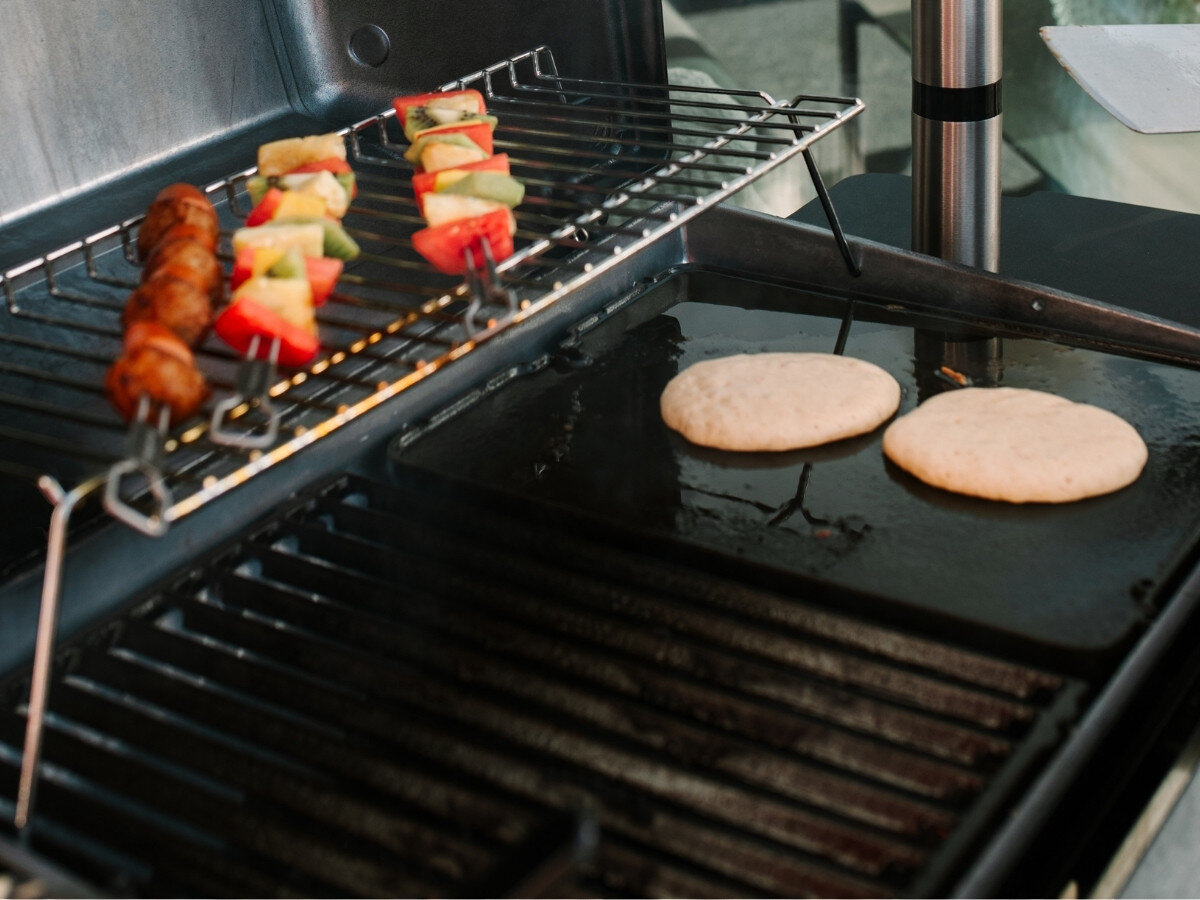 Płyta do grilla żeliwna BROIL KING GEM PORTA-CHEF 11237 33.5 x 26.5 cm, gładka powierzchnia, ryby, owoce morza, naleśniki, Osoba smaży na żeliwnej płycie grillowej naleśniki na gazowym grillu, obok na górnym ruszcie leżą kolorowe szaszłyki z warzyw i owoców.