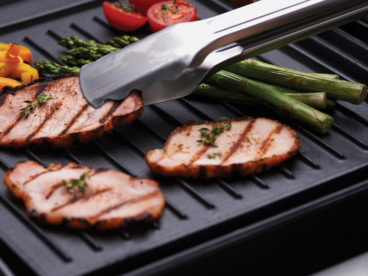 Płyta do grilla żeliwna BROIL KING GEM PORTA-CHEF 11237 33.5 x 26.5 cm, ryflowana płyta, mięso, grillowy wzór, warzywa, Kawałki grillowanego mięsa i zielonych szparagów leżą na ryflowanej płycie żeliwnej, chwytane stalowymi szczypcami.