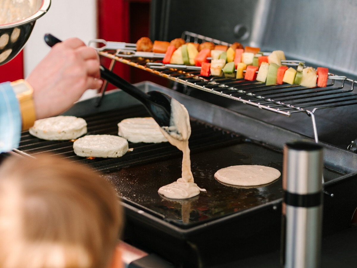 Płyta do grilla żeliwna BROIL KING GEM PORTA-CHEF 11237 33.5 x 26.5 cm, dopasowanie, grill Broil King, ruszt, stabilność, Zbliżenie na nalewanie ciasta naleśnikowego łyżką na rozgrzaną, gładką płytę żeliwną na grillu, w tle widać szaszłyki z warzyw i owoców.