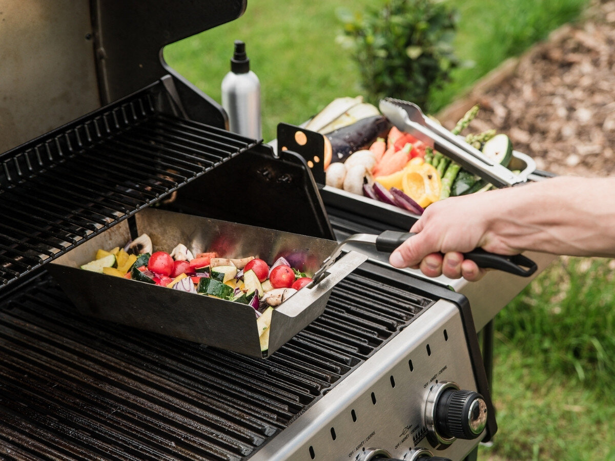Osoba trzyma uchwyt wielofunkcyjny do rusztów z naczyniem pełnym kolorowych warzyw na grillu. W tle widoczne są inne warzywa i akcesoria do grillowania.