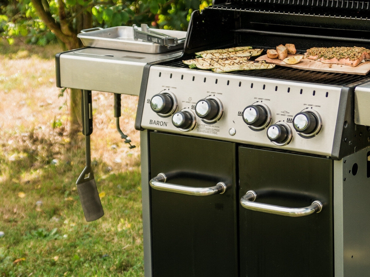 Na zdjęciu widoczny jest grill z czterema pokrętłami i napisem \'BARON\' na panelu. Na ruszcie znajdują się kawałki jedzenia, a obok wisi narzędzie kuchenne.
