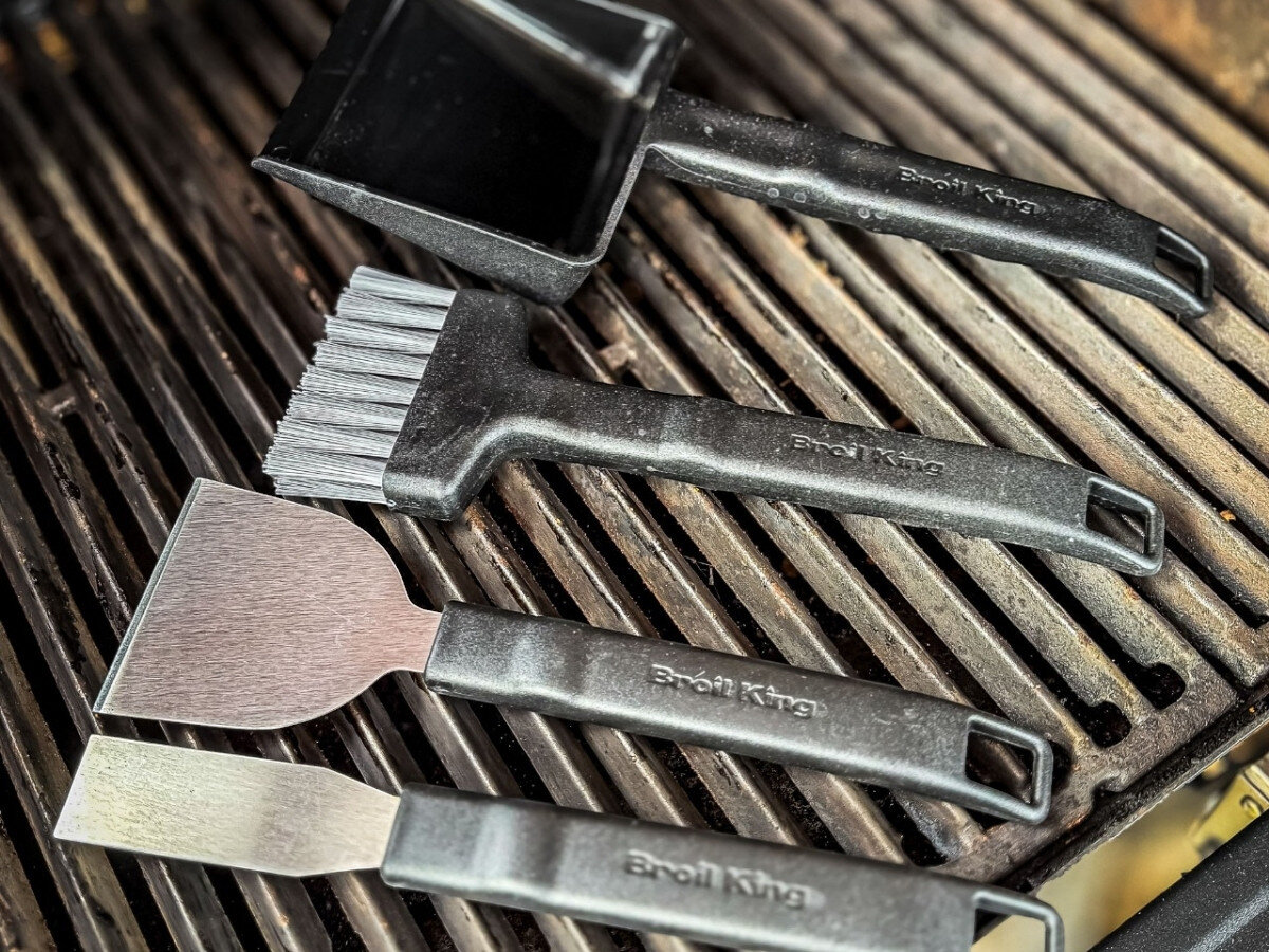 Na obrazie widoczny jest zestaw akcesoriów do czyszczenia grilla, w skład którego wchodzą szczotki i skrobaki z czarnymi uchwytami. Na uchwytach widnieje napis \'Broil King\'.