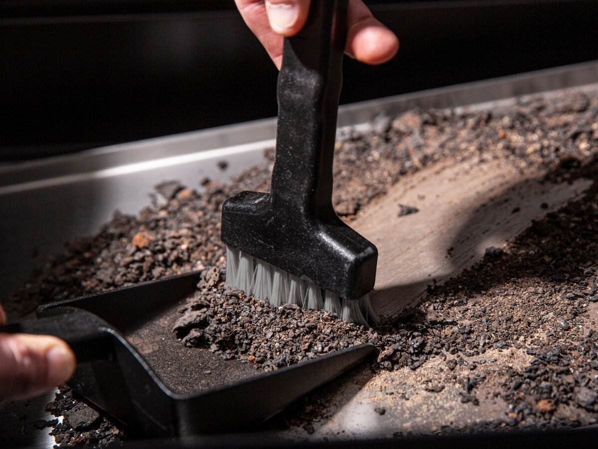 Osoba używa czarnej szczotki do czyszczenia powierzchni z resztek jedzenia i popiołu. Obok znajduje się czarna szufelka zbierająca zanieczyszczenia.