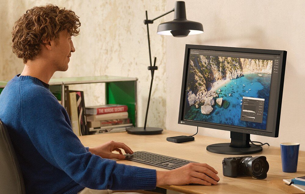 Monitor EIZO ColorEdge CS2400R Mężczyzna w niebieskim swetrze siedzi przy biurku i pracuje na komputerze z podłączonym monitorem. Na ekranie widoczne jest zdjęcie nadmorskiego klifu z błękitną wodą. Na biurku obok leży aparat fotograficzny, opis
