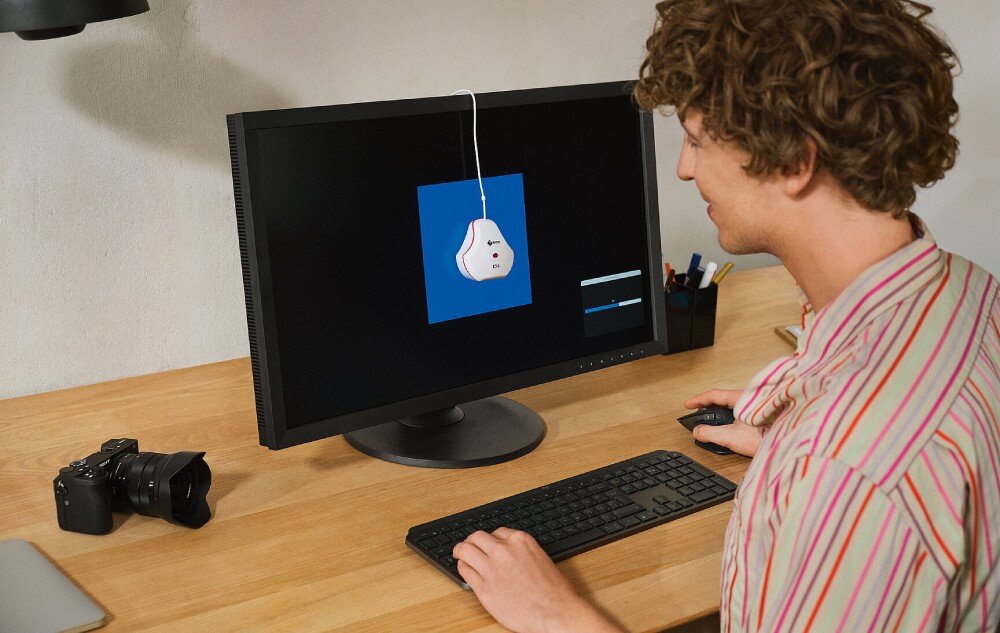 Monitor EIZO ColorEdge CS2400R Mężczyzna w koszulowej bluzce kalibruje monitor przy pomocy urządzenia zawieszonego na ekranie. Na biurku obok znajduje się aparat fotograficzny i klawiatura, kalibracja sprzętowa