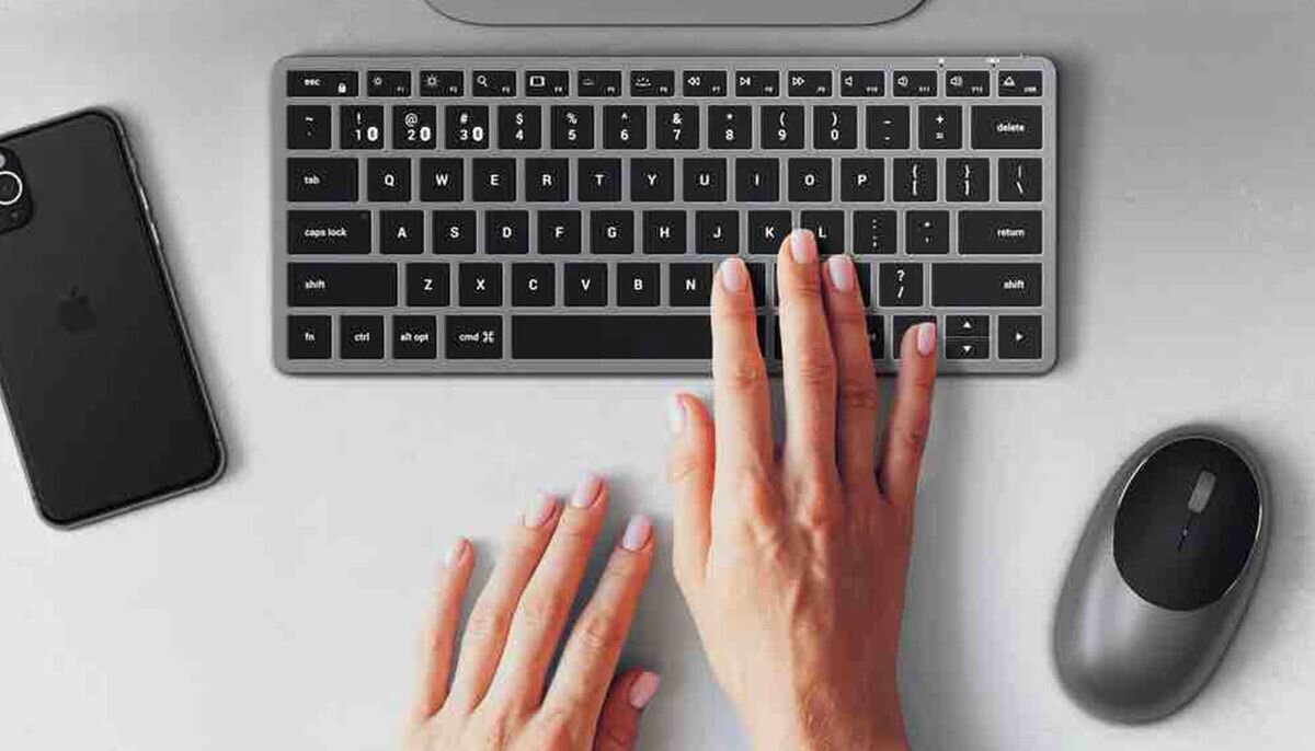 SATECHI Slim X1 Satechi Slim X1 bezprzewodowa klawiatura Bluetooth podświetlanymi klawiszami - klawiatura z myszką na blacie, obok smartfon, na klawiaturze kobiece dłonie
