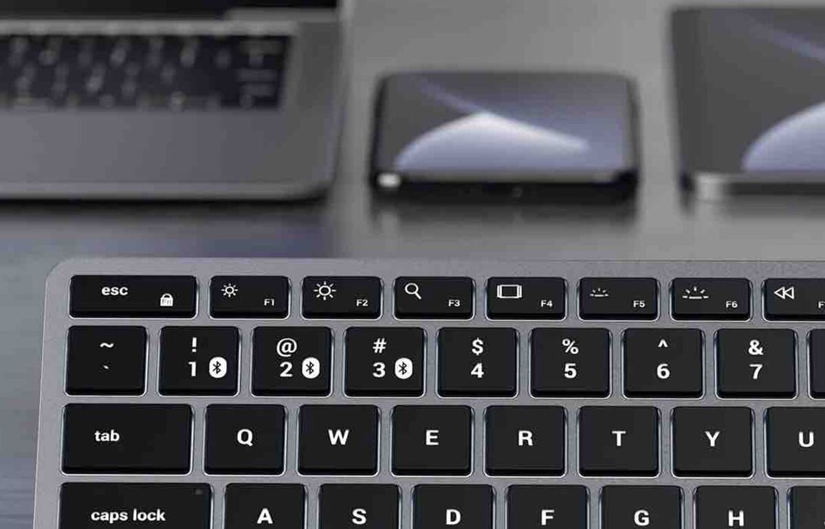 SATECHI Slim X1 technologia Bluetooth 5.0 jednoczesne sparowanie z kilkoma sprzętami - klawiatura na blacie, obok telefon i długopis - zbliżenie na klawisze, w tle widoczny laptop i smartfon