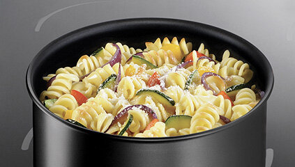 Czarny garnek wypełniony makaronem świderki z kawałkami warzyw, takich jak cukinia, papryka i cebula, posypany serem. Tło jest szare z delikatnymi, jasnymi liniami.