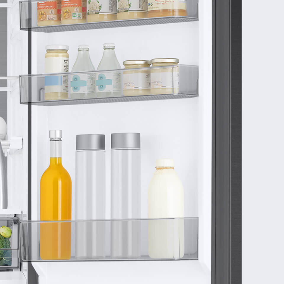 Wnętrze lodówki z trzema półkami na drzwiach, na których znajdują się różne produkty spożywcze, w tym butelki i słoiki. Na dolnej półce widoczne są dwie srebrne butelki, żółta butelka oraz biała butelka.