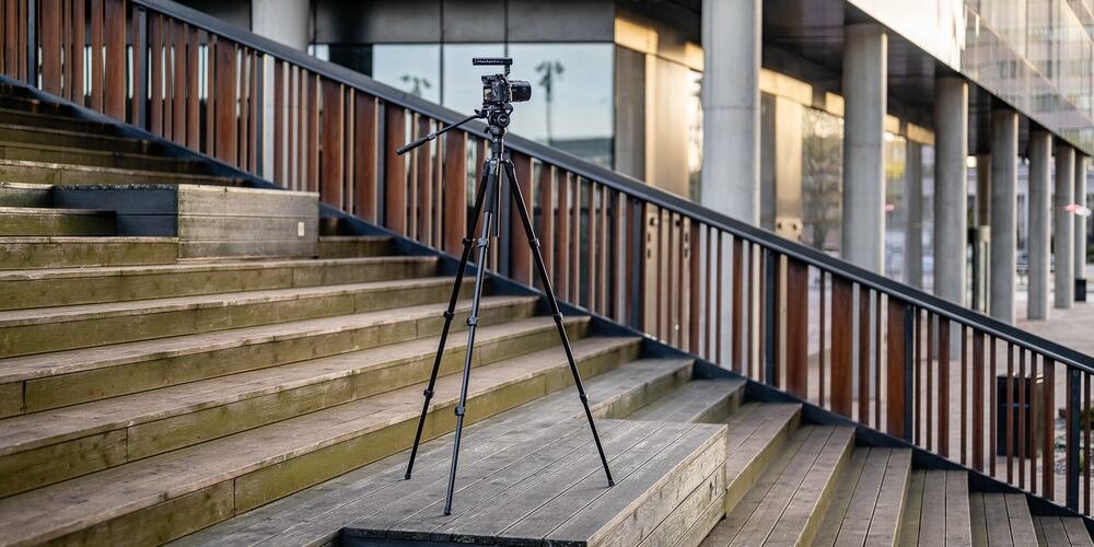 Aparat fotograficzny zamontowany na statywie Fotopro S5i PRO, ustawiony na drewnianych schodach na tle nowoczesnego budynku. kv początek opisu