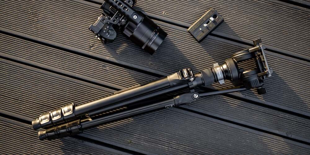 Na czarnych deskach kompozytowych leżą złożony statyw Fotopro S5I Pro z głowicą, płytka szybkiego montażu i lustrzanka.