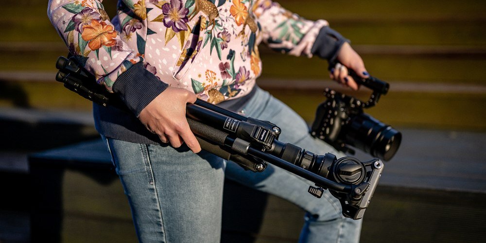 Zbliżenie na złożony statyw Fotopro S5i Pro niesiony w ręku przez kobietę. W drugiej ręce trzyma ona aparat na gimbalu.