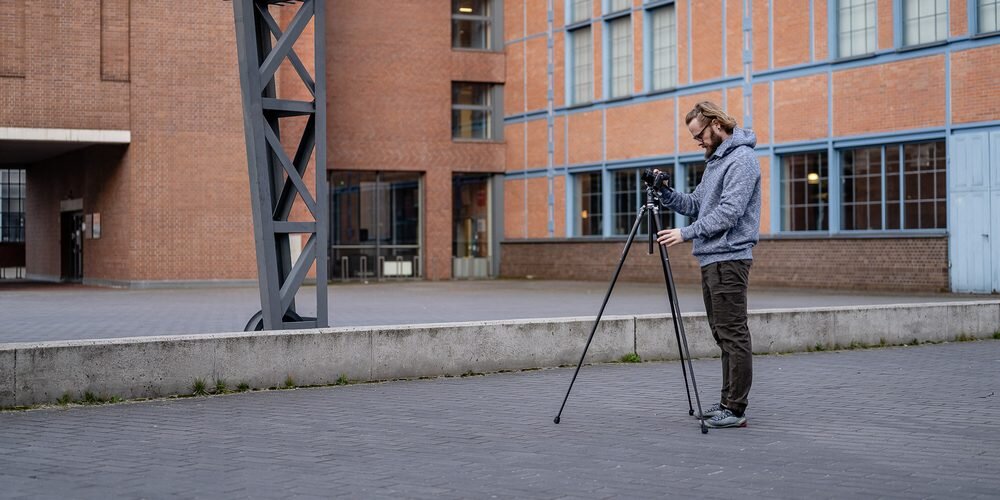 Osoba korzystająca z karbonowego statywu Fotopro X-Aircross 3 na zewnątrz, ustawiająca aparat na tle budynku z cegły.