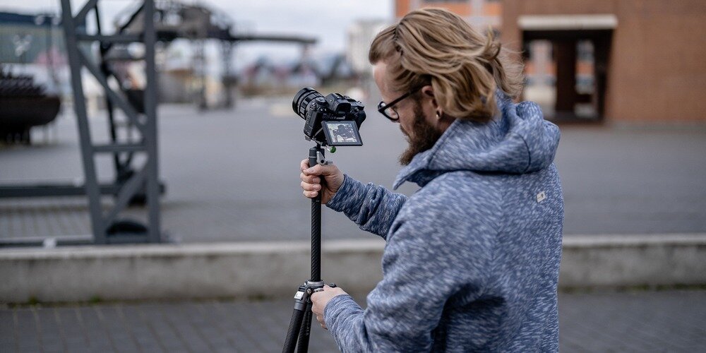 Fotograf obsługujący aparat zamocowany na statywie Fotopro X-Aircross 3 podczas pracy w plenerze na tle miejskiej architektury.