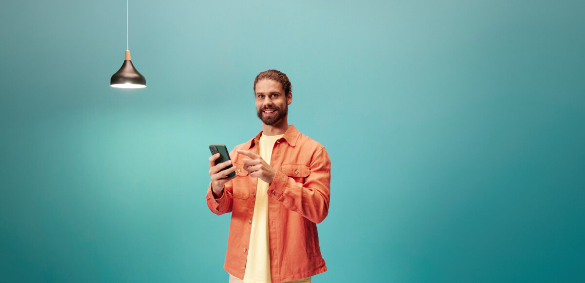 Wtyczka WIZ Smart 929002427201, energia, monitorowanie, śledzenie, aplikacja, na lazurowym tle mężczyzna obsługuje telefon, obok wisi włączona lampa sufitowa