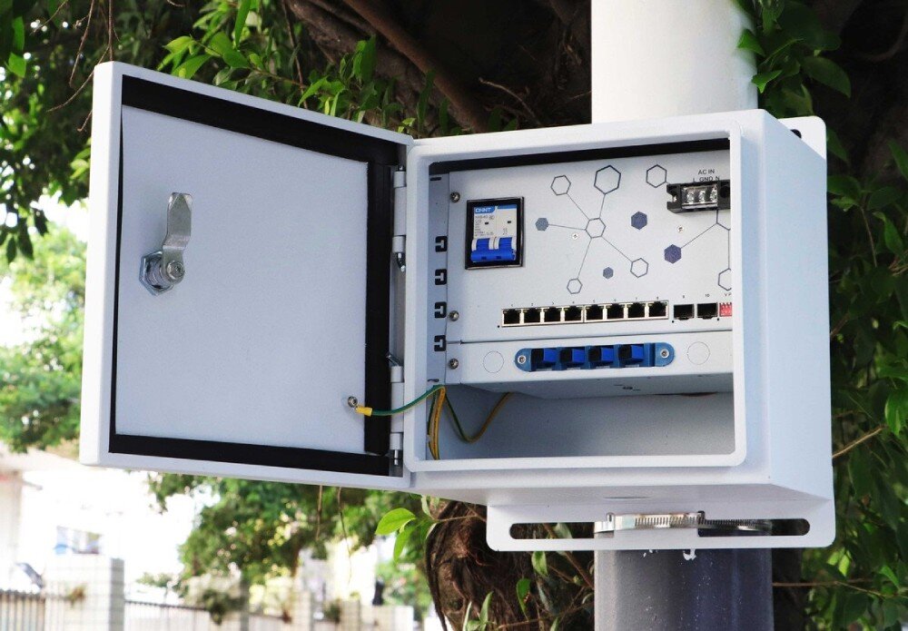 Switch EXTRALINK Atlas Otwarta biała obudowa zamontowana na słupie w otoczeniu roślinności ukazująca wewnętrzny panel z ośmioma portami ethernetowymi, dwoma gniazdami SFP oraz bezpiecznikiem automatycznym