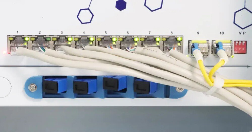 Switch EXTRALINK Atlas Detal panelu czołowego z wpiętymi szarymi przewodami sieciowymi i żółtymi światłowodami przy których widoczne są zapalone zielone lampki kontrolne